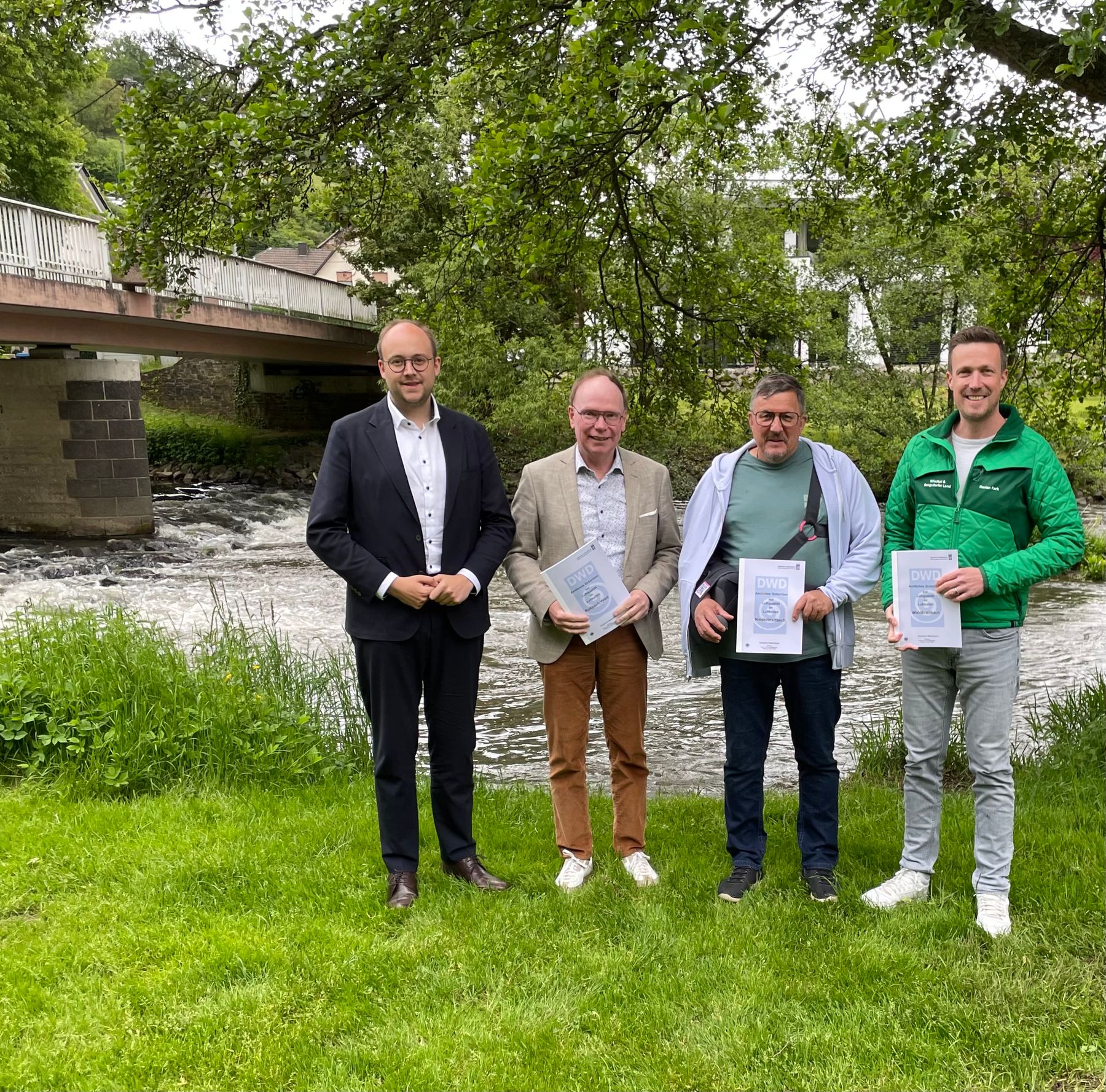 Four men stand on grass near a river, holding certificates and smiling. A bridge, trees, and houses are visible in the background, creating a lush, outdoor setting.